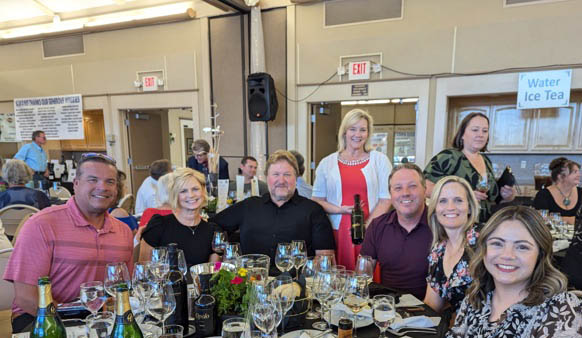 A group of people sitting at a round banquet table while one person is presenting a bottle of wine in the middle