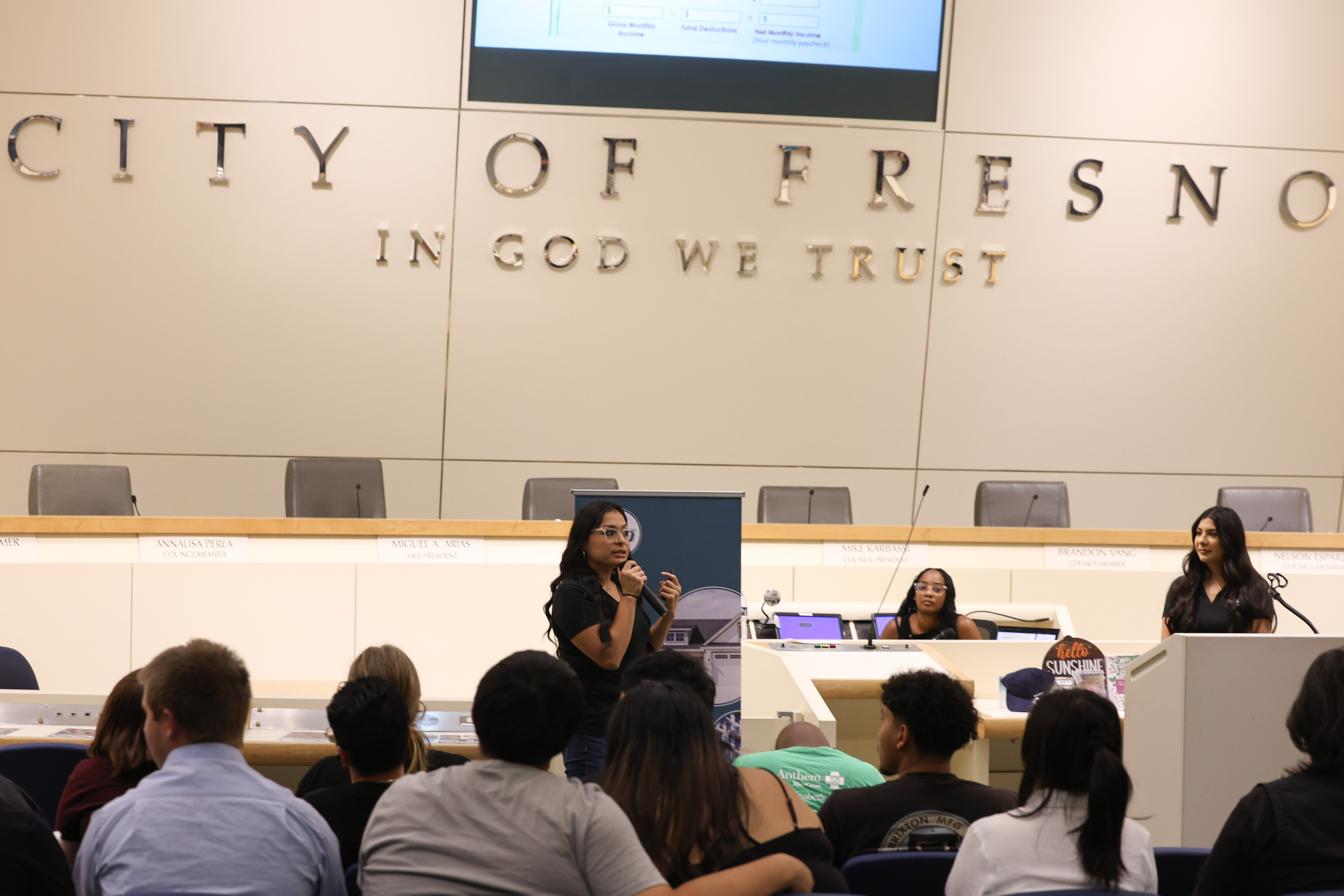 Two EECU employees presenting in front of an audience of youths, inside in the Council Chambers at Fresno City Hall.