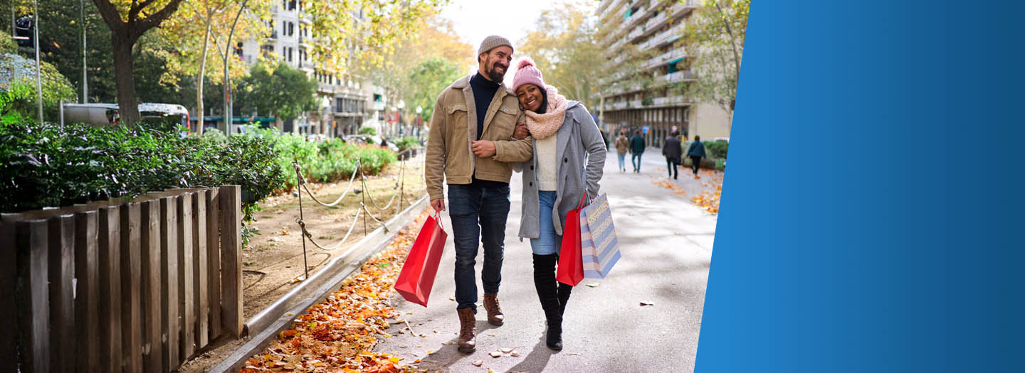 A man and woman smiling while walking down a street with people in the background, they are holding gift bags with a holiday theme