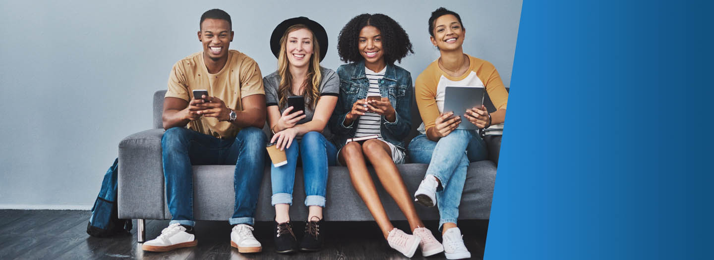 One young adult male and three young adult females sitting on a gray couch holding various technology devices